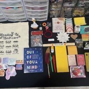 May include: A collection of stationery and lifestyle items including a canvas tote bag with the text Reading Is Self-care, a blue book titled Out Of Your Mind, various stickers, notebooks, pens, a yellow notepad, a yellow scrunchie, and small packets.