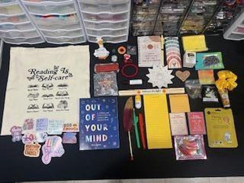 May include: A collection of stationery and lifestyle items including a canvas tote bag with the text Reading Is Self-care, a blue book titled Out Of Your Mind, various stickers, notebooks, pens, a yellow notepad, a yellow scrunchie, and small packets.