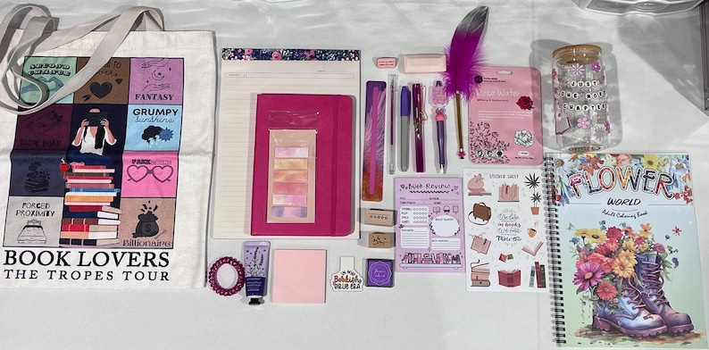 May include: A collection of stationery and book-themed items including a white tote bag with the text Book Lovers The Tropes Tour, a pink notebook, a spiral-bound Flower World coloring book, pens, a purple feather pen, stickers, and a glass jar.