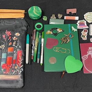 May include: A collection of stationery items including a black zippered pouch with a floral book print, a green notebook, a maroon journal with a bottle graphic, various pens, stickers, a green scrunchie, a small round container, and assorted metal charms.