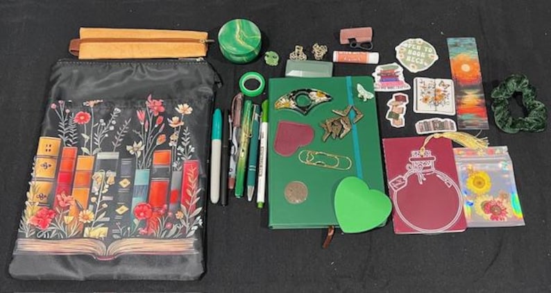 May include: A collection of stationery items including a black zippered pouch with a floral book print, a green notebook, a maroon journal with a bottle graphic, various pens, stickers, a green scrunchie, a small round container, and assorted metal charms.