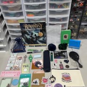 May include: A collection of book-themed items including a Romantasy coloring book, a green notebook, a beige pencil case with the text 'Don't Ask Me What I'm Reading', various stickers, bookmarks, a blue pouch, a black hand mirror, and pens.