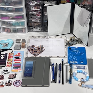 May include: A collection of stationery items including a gray notebook, a weekly planner, various pens, a heart-shaped notepad, stickers, bookmarks, a blue collagen face mask, a key-shaped pen, and a blue and white beaded keychain with a tassel.