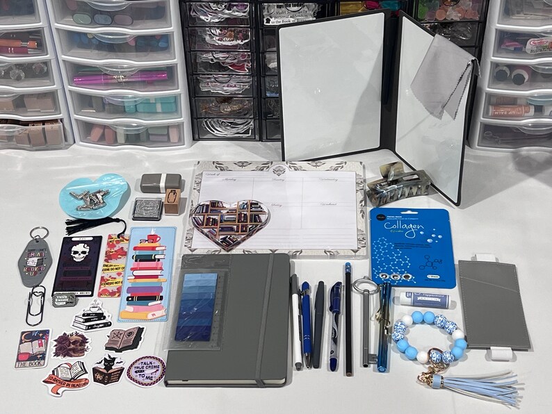 May include: A collection of stationery items including a gray notebook, a weekly planner, various pens, a heart-shaped notepad, stickers, bookmarks, a blue collagen face mask, a key-shaped pen, and a blue and white beaded keychain with a tassel.
