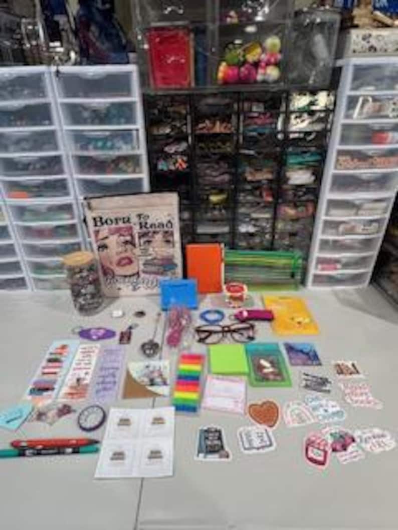 May include: A collection of stationery and reading accessories including a pouch with the text Born To Read, a glass jar, an orange notebook, reading glasses, various bookmarks, colorful sticky notes, pens, and numerous decorative stickers.
