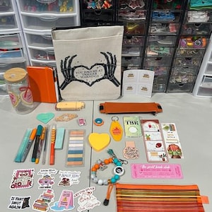 May include: A collection of stationery and book-themed accessories including a canvas pouch with skeleton hands forming a heart and the text 'YOU HAD ME AT TRIGGER WARNINGS', a glass tumbler, stickers, pens, bookmarks, a beaded bracelet, and a mesh pencil case.