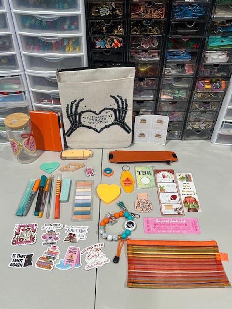 May include: A collection of stationery and book-themed accessories including a canvas pouch with skeleton hands forming a heart and the text 'YOU HAD ME AT TRIGGER WARNINGS', a glass tumbler, stickers, pens, bookmarks, a beaded bracelet, and a mesh pencil case.