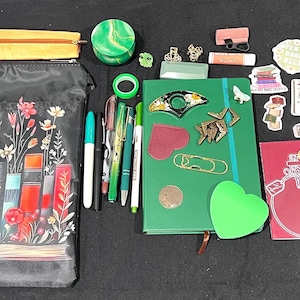 May include: A collection of stationery items including a black zippered pouch with a floral book print, a green notebook, various pens, a yellow pencil case, stickers, a bookmark, a green scrunchie, a small green container, and assorted paper clips.