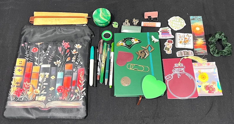 May include: A collection of stationery items including a black zippered pouch with a floral book print, a green notebook, various pens, a yellow pencil case, stickers, a bookmark, a green scrunchie, a small green container, and assorted paper clips.