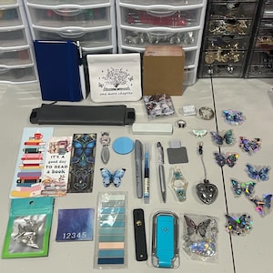 May include: A collection of stationery and craft items including a blue notebook, a white pouch with a book illustration, bookmarks, pens, butterfly-shaped stickers, a blue light, a heart-shaped charm, and various small accessories arranged on a surface.