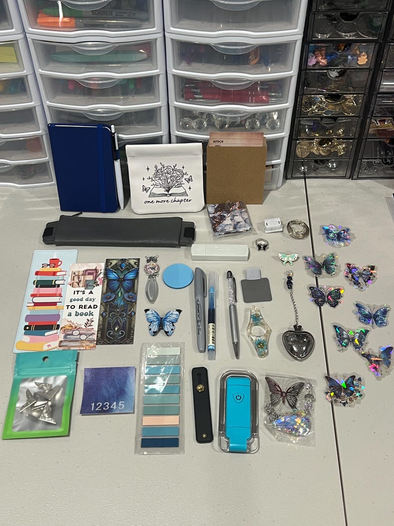 May include: A collection of stationery and craft items including a blue notebook, a white pouch with a book illustration, bookmarks, pens, butterfly-shaped stickers, a blue light, a heart-shaped charm, and various small accessories arranged on a surface.