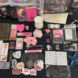 May include: A collection of stationery and lifestyle items including a pink notebook, a white wax warmer, various stickers, bookmarks, a pink scrunchie, a star-shaped dish, a small mirror, pens, and a canvas pouch with the text artfullyegg printed on it.