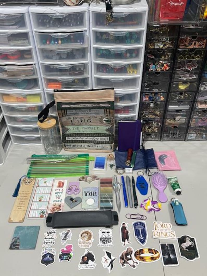 May include: A collection of items including a canvas tote bag with book titles, a glass jar, a purple notebook, a green pencil case, various bookmarks, pens, stickers featuring fantasy themes, a hairbrush, a small tube, and a black pencil case.