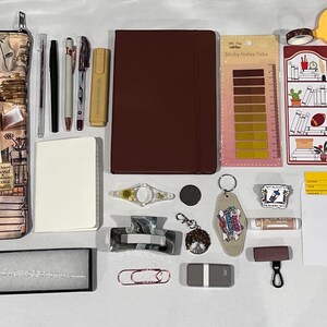 May include: A collection of stationery and accessories including a book-themed zippered pouch, a dark red notebook, a white notepad, various pens, a glass dip pen with ink, sticky index tabs, bookmarks, stickers, a hair clip, and a gold eye mask.