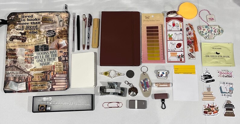 May include: A collection of stationery and accessories including a book-themed zippered pouch, a dark red notebook, a white notepad, various pens, a glass dip pen with ink, sticky index tabs, bookmarks, stickers, a hair clip, and a gold eye mask.