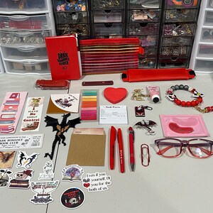 May include: A collection of red-themed stationery and accessories including a notebook labeled Dark Romance Reader, pens, stickers with dragon and book motifs, a heart-shaped notepad, a beaded bracelet, red-rimmed glasses, and various small trinkets.