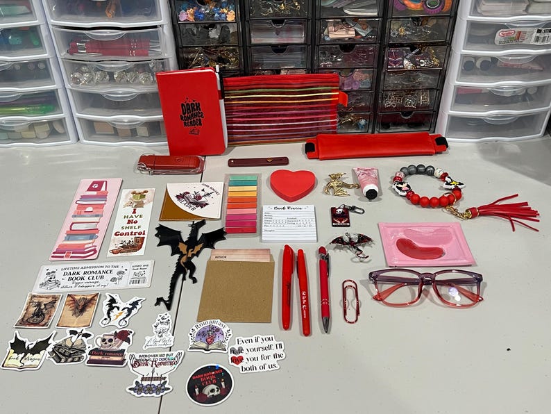 May include: A collection of red-themed stationery and accessories including a notebook labeled Dark Romance Reader, pens, stickers with dragon and book motifs, a heart-shaped notepad, a beaded bracelet, red-rimmed glasses, and various small trinkets.