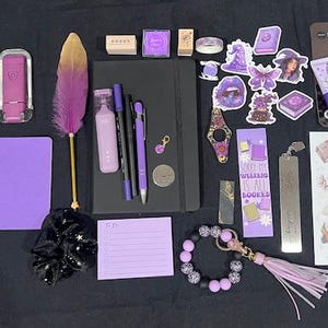 May include: A collection of stationery items including a black notebook, a purple feather pen, various purple pens, a charcoal face mask, a sticker sheet, a purple notepad, a beaded keychain, a scrunchie, and several purple-themed decorative stickers.
