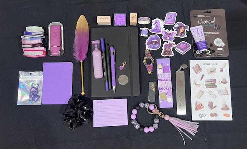 May include: A collection of stationery items including a black notebook, a purple feather pen, various purple pens, a charcoal face mask, a sticker sheet, a purple notepad, a beaded keychain, a scrunchie, and several purple-themed decorative stickers.