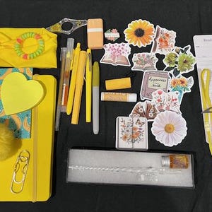 May include: A collection of yellow stationery and accessories including a headband, a notebook with a heart sticky note, a pom-pom, pens, markers, a lip balm, floral stickers, a glass dip pen in a box, and two printed book tracker bookmarks.