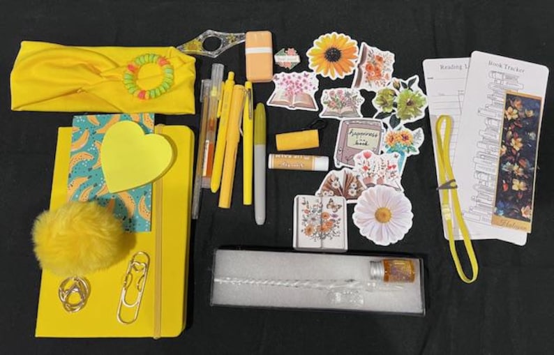 May include: A collection of yellow stationery and accessories including a headband, a notebook with a heart sticky note, a pom-pom, pens, markers, a lip balm, floral stickers, a glass dip pen in a box, and two printed book tracker bookmarks.