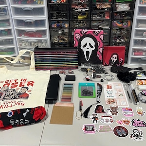 May include: A cream-colored canvas tote bag with a red and black horror-themed graphic and the text "GET IN LOSER WE'RE GOING KILLING," accompanied by a collection of stationery, stickers, a Ghostface notebook, a pencil case, and accessories.