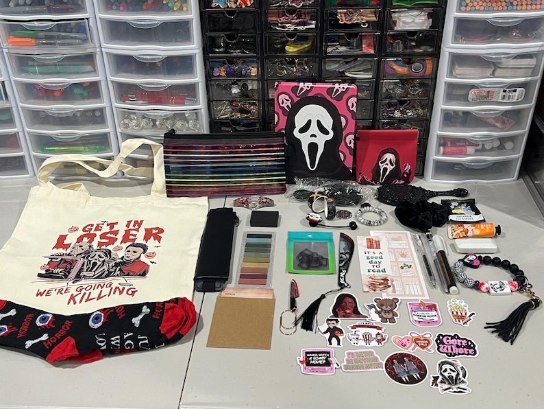 May include: A cream-colored canvas tote bag with a red and black horror-themed graphic and the text "GET IN LOSER WE'RE GOING KILLING," accompanied by a collection of stationery, stickers, a Ghostface notebook, a pencil case, and accessories.
