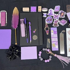 May include: A collection of stationery and accessories including a black notebook, a purple feather quill, various purple pens, a charcoal face mask, a sticker sheet, a purple notepad, a beaded keychain, a black scrunchie, and assorted purple stickers.