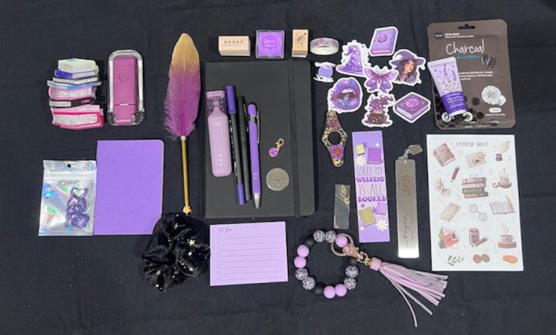 May include: A collection of stationery and accessories including a black notebook, a purple feather quill, various purple pens, a charcoal face mask, a sticker sheet, a purple notepad, a beaded keychain, a black scrunchie, and assorted purple stickers.