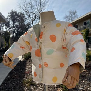 May include: A reversible, button-up jacket with a cream-colored exterior adorned with orange, yellow, and teal circular patterns. The jacket's interior is a light green color. The jacket is displayed on a mannequin.