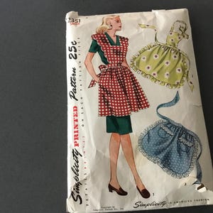 May include: Vintage Simplicity pattern 2351 for aprons. The pattern envelope features a woman in a red and white checkered apron, green dress, and brown shoes. Additional apron designs are shown in the image. The text "Simplicity Printed Pattern" is visible.