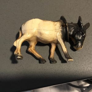 May include: A small, vintage donkey figurine. The toy donkey is painted with a cream-colored body and a black head and legs. It has a harness around its neck and is posed in a walking position.