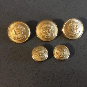 May include: Five gold-colored buttons with a raised design featuring a crown and a crest. The buttons are different sizes.