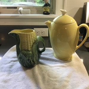 May include: A yellow ceramic teapot with a lid and a green ceramic pitcher shaped like an ear of corn.