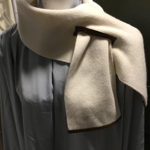 May include: Cream-colored scarf with a ribbed knit texture and a dark brown leather accent. The scarf is draped over a light blue garment, showcasing its design and texture. The scarf appears to be made of a soft, warm material, suitable for cooler weather.