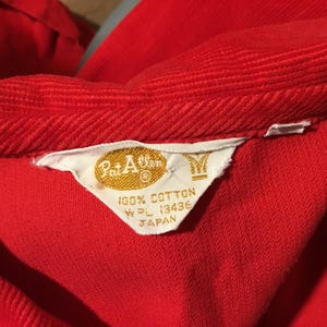 K&ouml;nnte beinhalten: Nahaufnahme eines roten Cordkleidungsst&uuml;cks, m&ouml;glicherweise ein Hemd oder eine Jacke, mit einem sichtbaren Etikett. Das Etikett, in Wei&szlig; und Gold, lautet "Pat Allen" und "100% Baumwolle". Die Textur und Farbe des Kleidungsst&uuml;cks sind deutlich sichtbar.