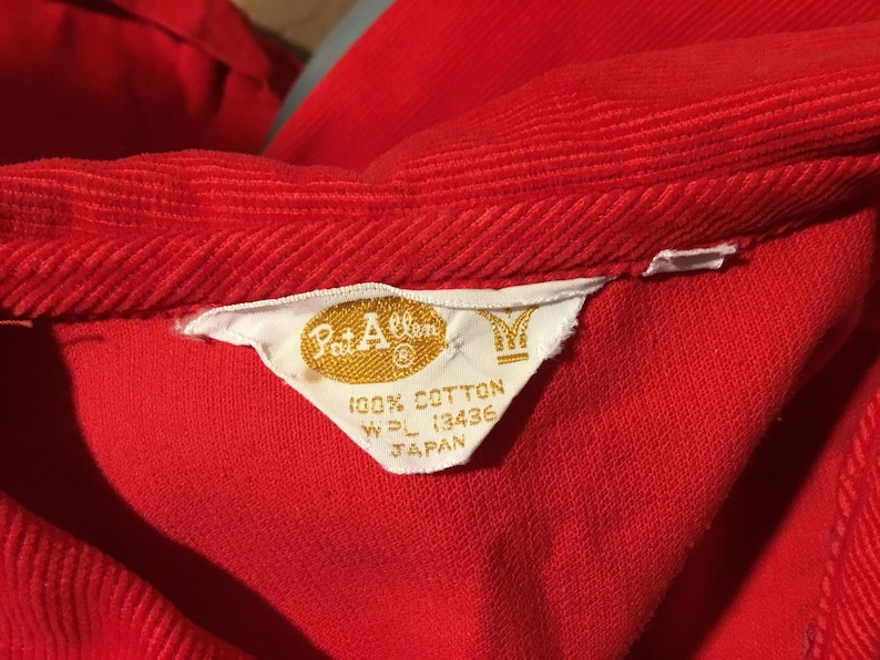 K&ouml;nnte beinhalten: Nahaufnahme eines roten Cordkleidungsst&uuml;cks, m&ouml;glicherweise ein Hemd oder eine Jacke, mit einem sichtbaren Etikett. Das Etikett, in Wei&szlig; und Gold, lautet "Pat Allen" und "100% Baumwolle". Die Textur und Farbe des Kleidungsst&uuml;cks sind deutlich sichtbar.