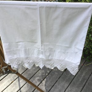 May include: White linen sheet with a decorative lace trim along the bottom edge, hanging on a wooden drying rack. The sheet is made of a smooth, opaque fabric. The lace trim adds a delicate, vintage touch.