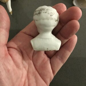 May include: A small, white porcelain doll head with a detailed hairstyle. The head is a bust, showing the neck and shoulders. The porcelain has some age-related discoloration.