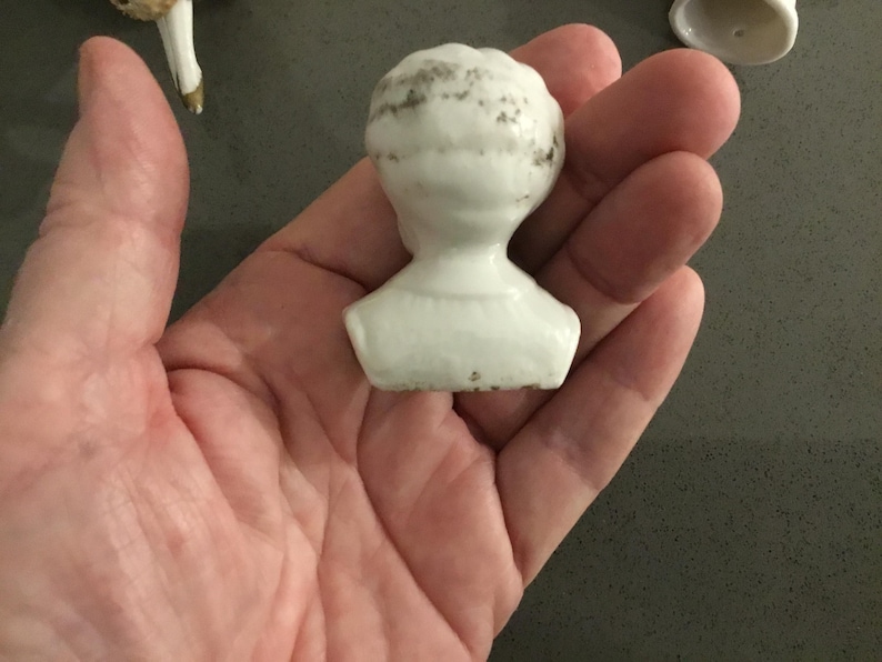 May include: A small, white porcelain doll head with a detailed hairstyle. The head is a bust, showing the neck and shoulders. The porcelain has some age-related discoloration.