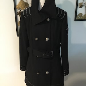 May include: A black double-breasted coat featuring a high collar, silver-toned buttons, and a matching belt with a buckle. The shoulders are accented with silver chain details, while the cuffs are embellished with small silver studs.