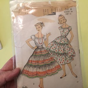 May include: Vintage sewing pattern packet featuring illustrations of two dresses. The dresses have tiered skirts and off-the-shoulder sleeves. The packet is in a clear plastic sleeve, with text indicating measurements: bust 28 inches, waist 24 inches, and hip 31 inches.