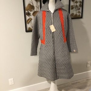 May include: A black and white gingham coat with orange trim. The coat has a zipper closure and long sleeves. The coat is on a white mannequin with a black beret.