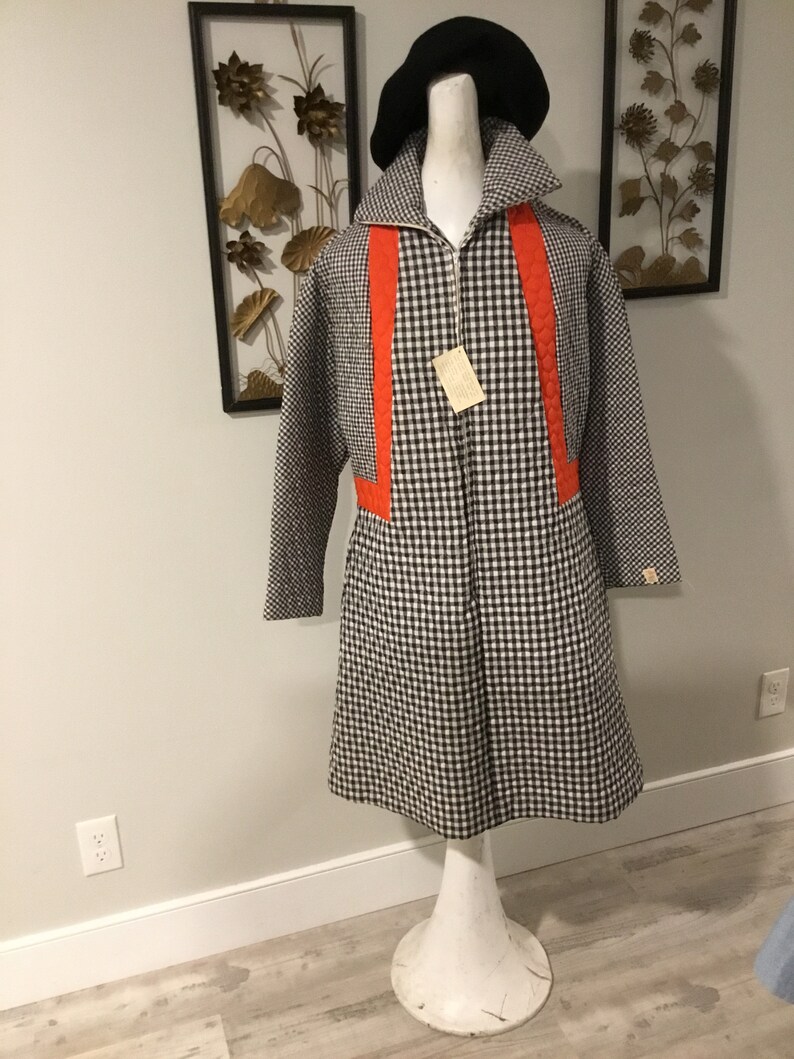 May include: A black and white gingham coat with orange trim. The coat has a zipper closure and long sleeves. The coat is on a white mannequin with a black beret.