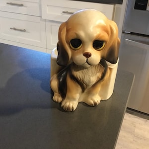 May include: A ceramic figurine of a brown and white dog with big, sad eyes. The dog is sitting in a white planter.