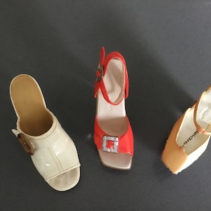 May include: Three pairs of women's shoes. One pair is white with a buckle, one pair is orange with a buckle, and one pair is beige with a strap.