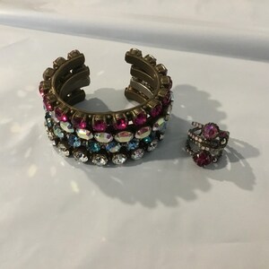 May include: A gold-coloured cuff bracelet and a ring adorned with colourful rhinestones. The bracelet has rows of pink, blue, and clear stones. The ring features a large pink stone surrounded by smaller stones. Jewellery.