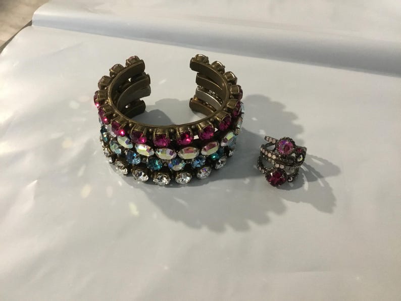 May include: A gold-coloured cuff bracelet and a ring adorned with colourful rhinestones. The bracelet has rows of pink, blue, and clear stones. The ring features a large pink stone surrounded by smaller stones. Jewellery.