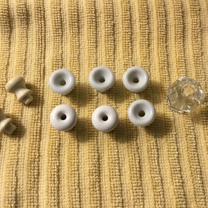 May include: A collection of white ceramic knobs and a clear glass knob. There are eight white ceramic knobs with a small hole in the centre of each. The glass knob is faceted and has a small hole in the centre.
