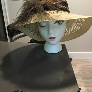 May include: A beige straw hat with a wide brim and a brown feather trim. The hat has a delicate crocheted design and a small brown bead accent.